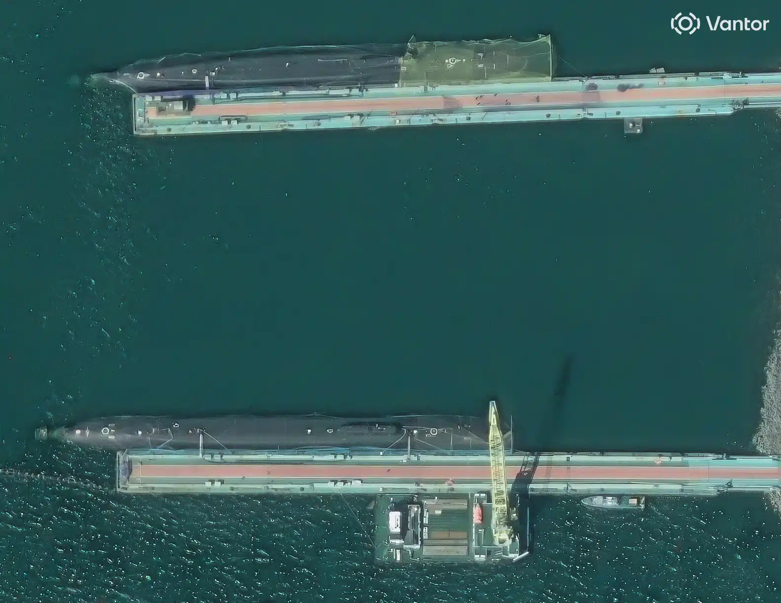 Satellite image of the Rybachiy base on Kamchatka: two large submarines alongside piers, covered along their full length by netting, one yellowish and one dark; a crane barge is working alongside