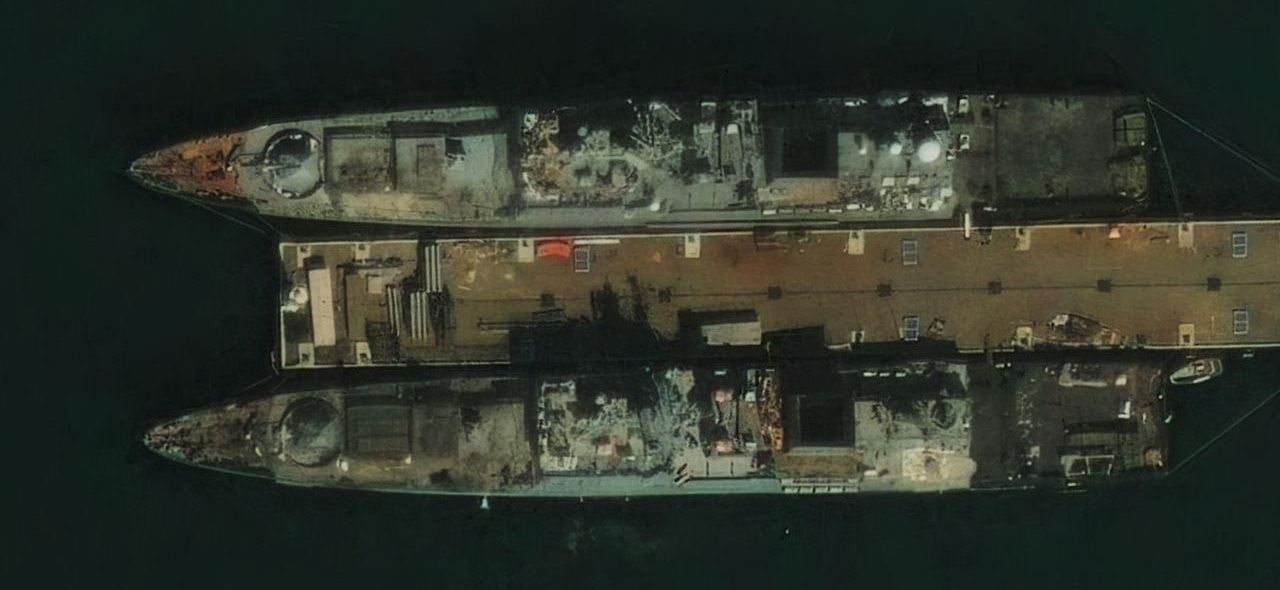 High-resolution satellite image showing two Project 11356R frigates lying parallel along opposite sides of a single pier in Novorossiysk, with dark soot patches visible on their decks